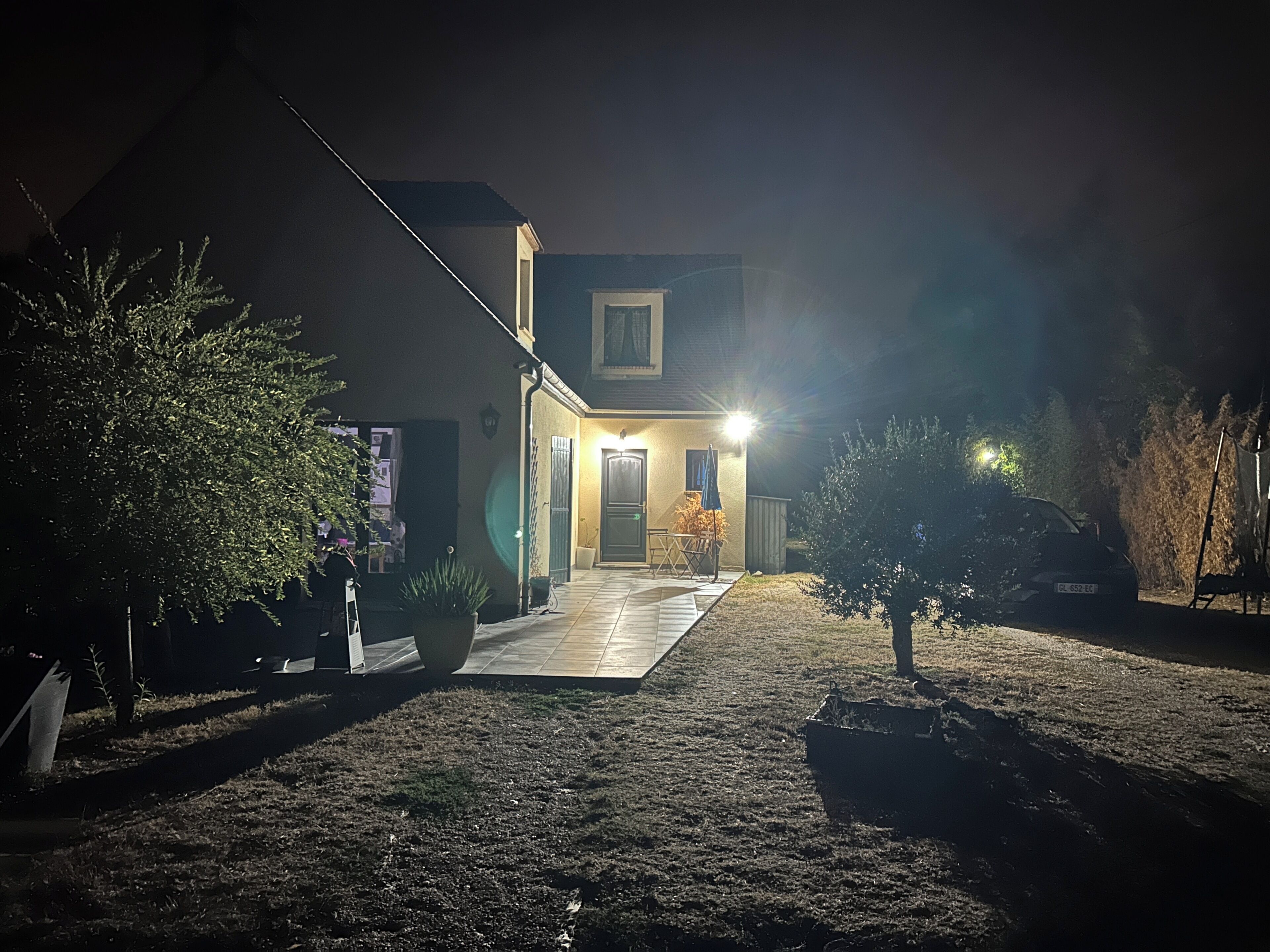 Ein Haus mit einem gepflasterten Einfahrtsweg, einem kleinen Garten mit einer Bank und einem geparkten Auto auf dem Weg.
