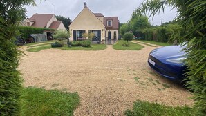 Una casa con vialetto in ghiaia, un'auto blu parcheggiata a destra e un prato curato con un piccolo albero.