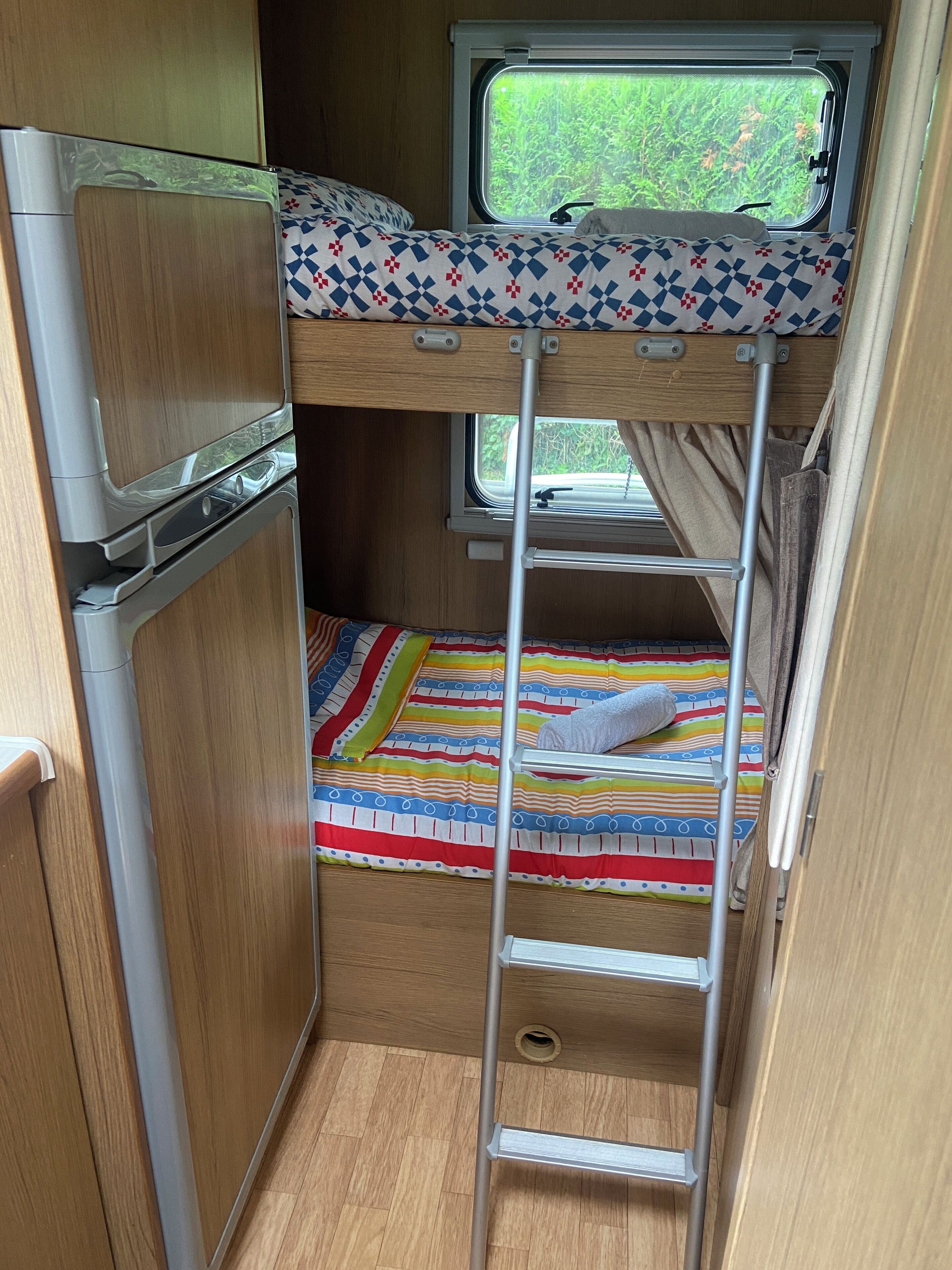 Interior d'una caravana compacta amb un llit superposat, una finestra petita que donava a zona verda i un espai de cuina petit.