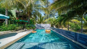 A swimming pool with two inflatable rings, surrounded by palm trees and a building with a green roof. - Holiday Villa 3BR Sea View An Bang Beach (Da Nang)