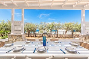 Un área de comedor al aire libre con mesas blancas, vasos de vidrio azules y un muro de piedra de fondo.