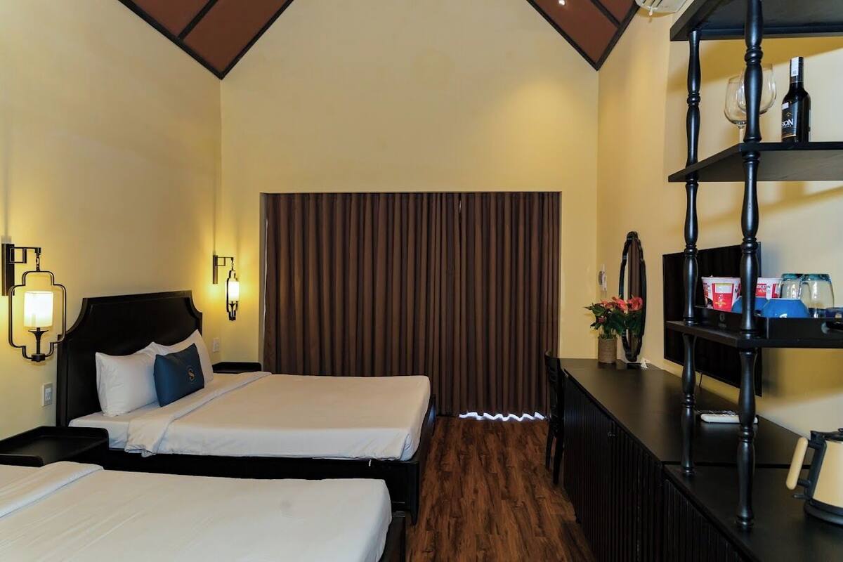 A hotel room with two beds, a wooden floor, a dark headboard, a wall-mounted shelf with bottles, and a curtain.