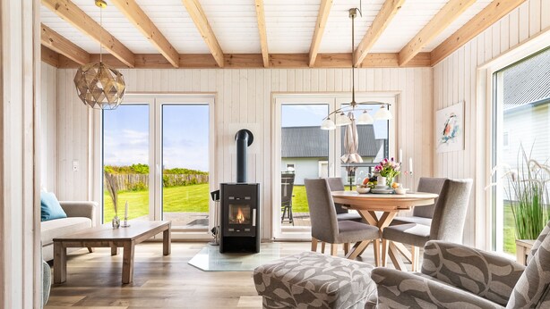 Una habitación acogedora con chimenea, vigas de madera en el techo, una mesa de comedor con sillas y ventanas amplias que ofrecen vistas al exterior.