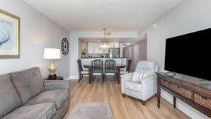 A modern living room with a gray sofa, a dining table with chairs, a TV on a wooden stand, and a kitchen in the background. - Oceanfront 3bed 3bath condo! 19th floor. Amazing views indoor & outdoor pools (Myrtle Beach)
