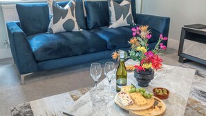 A living room with a blue sofa, a marble coffee table with a cheese board, wine glasses, and a bottle of wine. - Elliot Oliver - Comfortable Five Bedroom Town Centre House With Garden & Garage (Cheltenham)