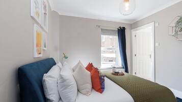 Un dormitorio con una cama, almohadas, una mesita de noche con lámpara, una ventana con cortinas y una obra de arte en la pared.
