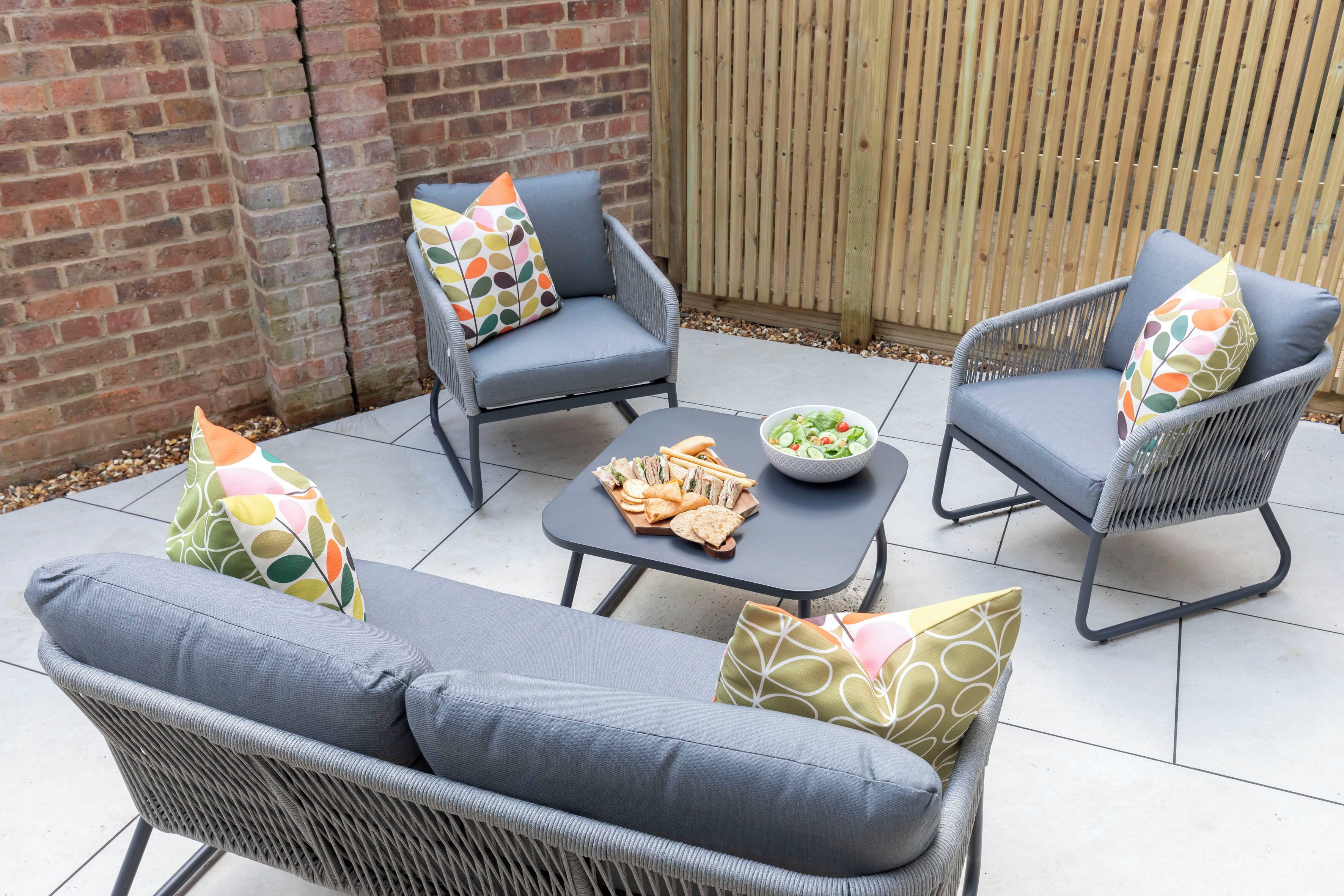 Een terras met rieten meubels en kussens, een tafel met lekkernijen, en een houten omheining op de achtergrond.