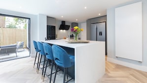A modern kitchen with a central island, bar stools, and a vase of flowers. - Elliot Oliver - Comfortable Five Bedroom Town Centre House With Garden & Garage (Cheltenham)