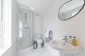 Un baño moderno con ducha envuelta en vidrio, inodoro blanco, lavamanos con artículos de aseo y un espejo grande.
