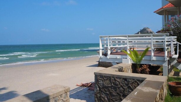 A beach with a white fence overlooking the ocean, a stone wall, and a building with a red-tiled roof. - Aranava Resort Huahin (Hua Hin)