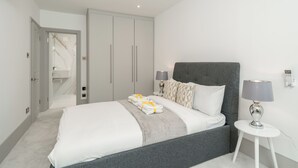 Et moderne soveværelse med en grå sengegavl, hvide sengetæpper, et natbord med en lampe og et garderobeskab.