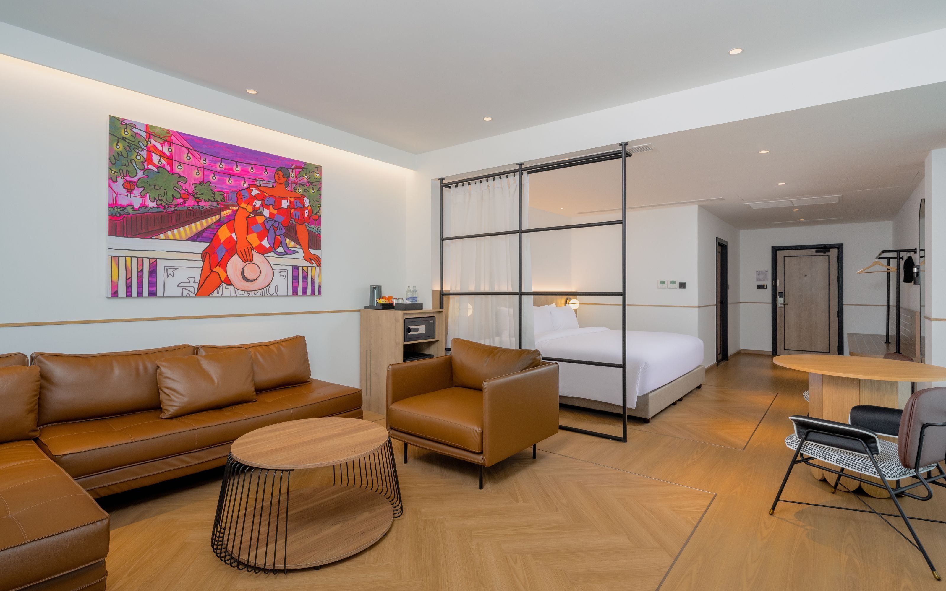 Una habitación de hotel moderna con un sofá de cuero marrón, una mesa de centro redonda de madera, una cama con sábanas blancas y un cuadro abstracto de colores en la pared.