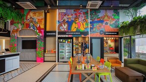 A vibrant hotel lobby with colorful murals, a central seating area with a wooden table, and a variety of amenities including a refrigerator, seating options, and a view of the outdoors. - Lub d Bangkok Chinatown (Bangkok)
