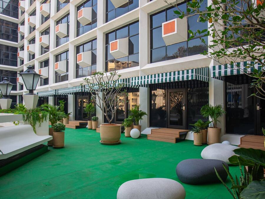 A modern building with a courtyard featuring green flooring, potted plants, outdoor seating, and a striped awning.