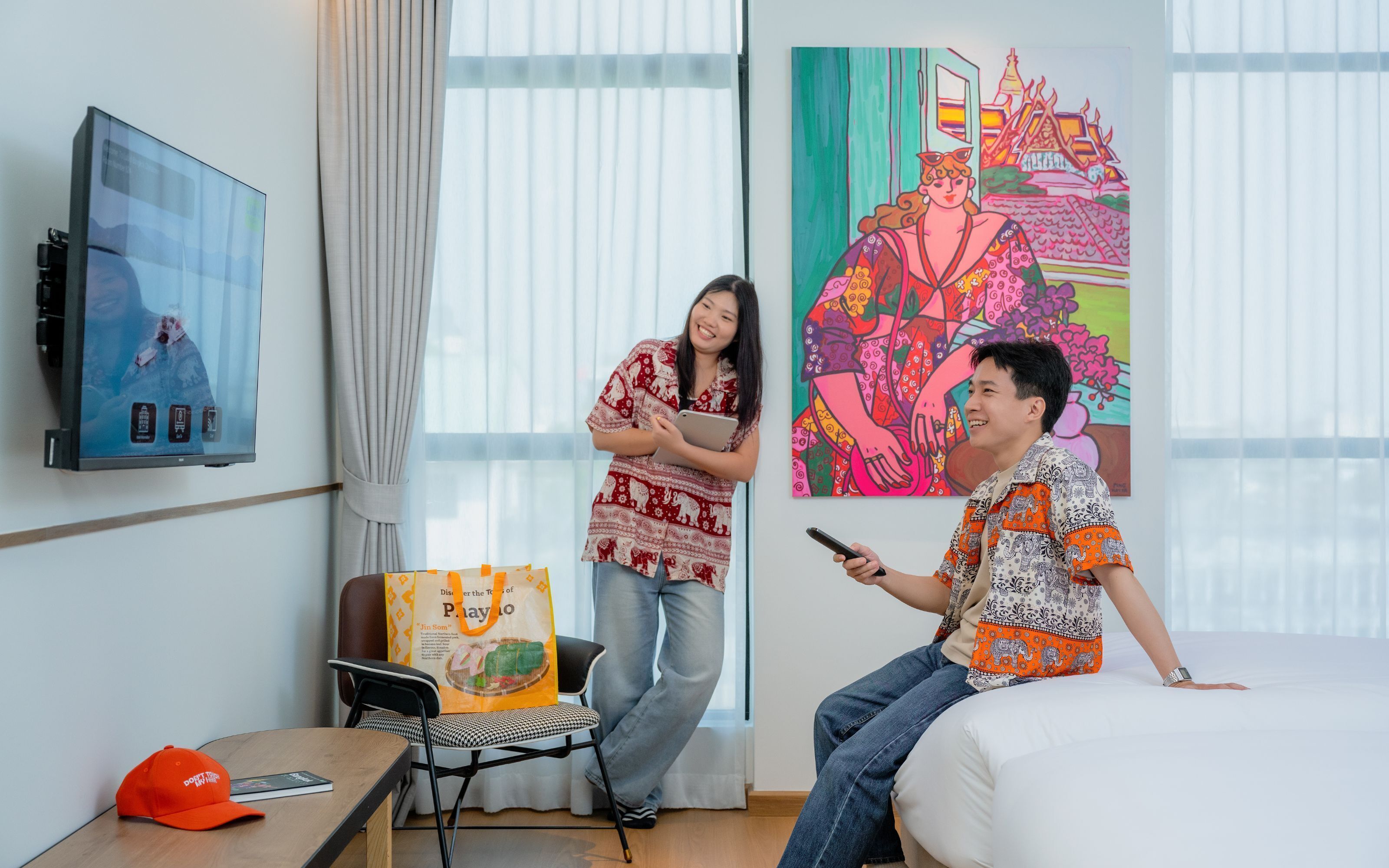 A hotel room with a bed, a desk with a laptop, a chair, a TV on the wall, and a colorful painting.