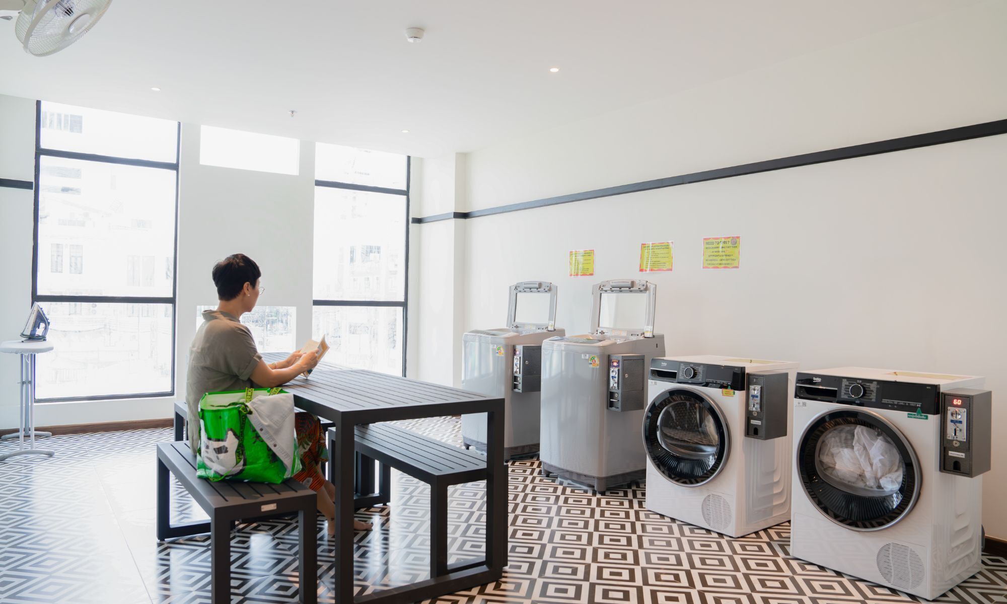 A laundry room with large windows, a table with a green bag, and multiple washing machines.