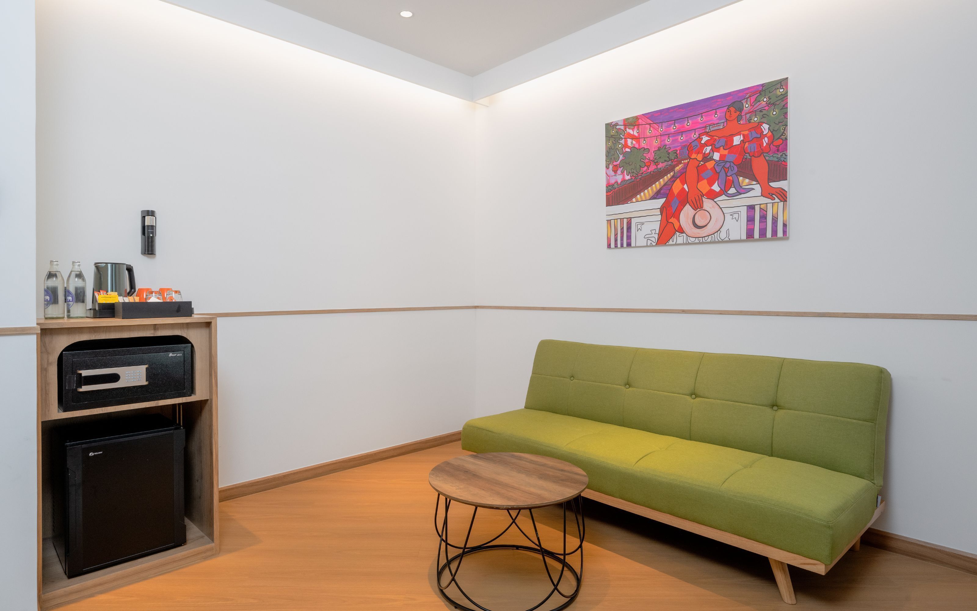 Habitación de hotel con sofá verde, una mesita de madera, un minibar y un cuadro en la pared.