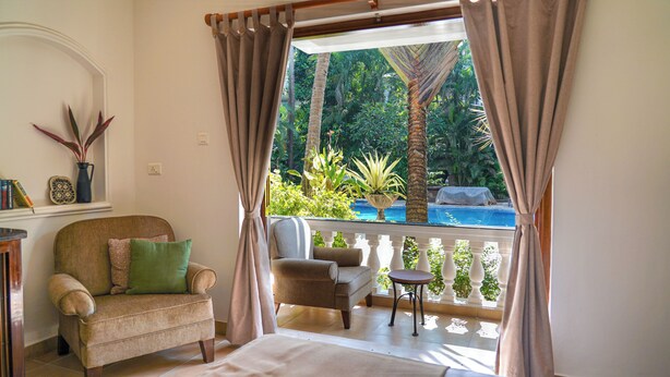 A room with a view of a pool, two chairs, a small table, and a sofa with a green pillow. - Olive Hotel Alidia Beach Resort by Embassy Group (Baga)