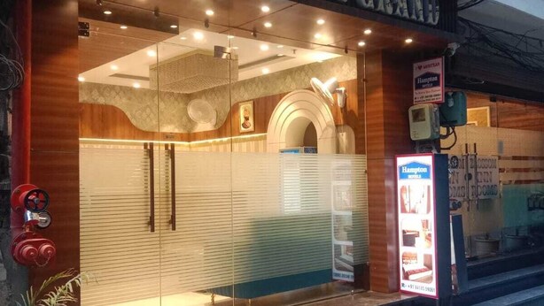 A hotel entrance with a modern design, featuring a glass facade, a wooden reception desk, and a visible sign for Hampton Hotels. - HOTEL JR GRAND (Amritsar)