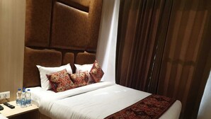 A hotel room with a large bed, a headboard, a bedside table with water bottles, and a window with curtains. - HOTEL JR GRAND (Amritsar)