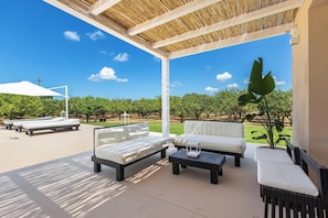 Um pátio externo com sofás brancos e mesas pretas, uma pérgula com cobertura de bambu e vista para um pomar.