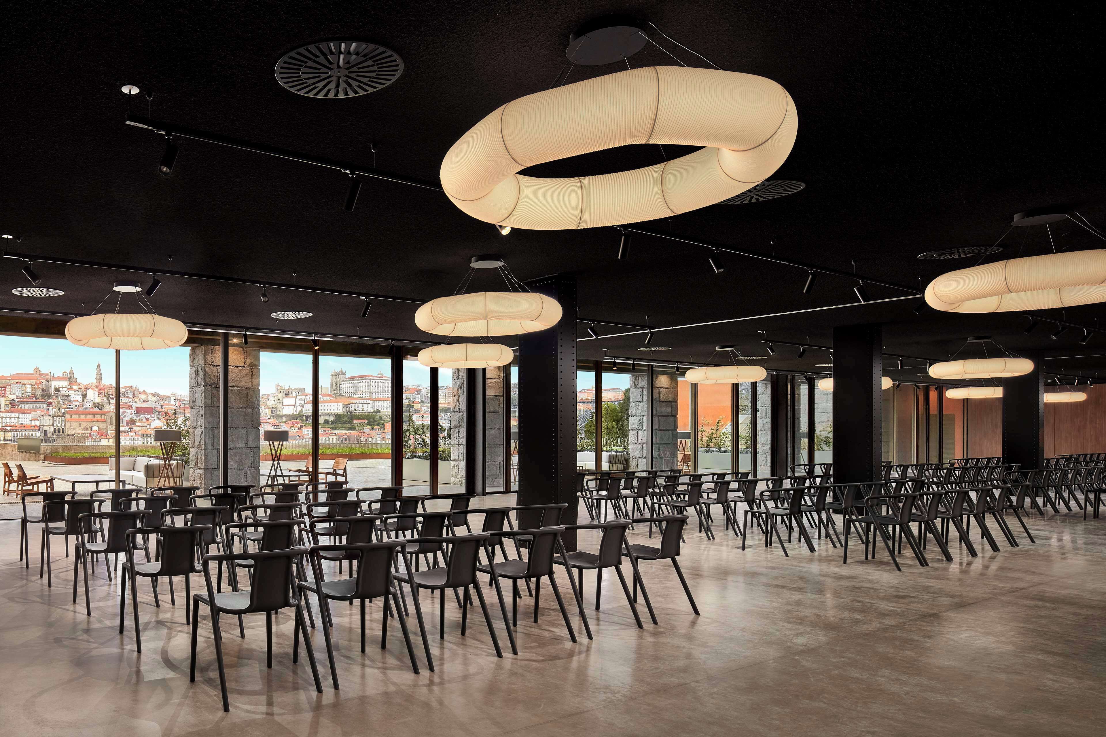 A spacious indoor area with rows of black chairs and tables, large circular hanging lights, and large windows offering a view of a cityscape.
