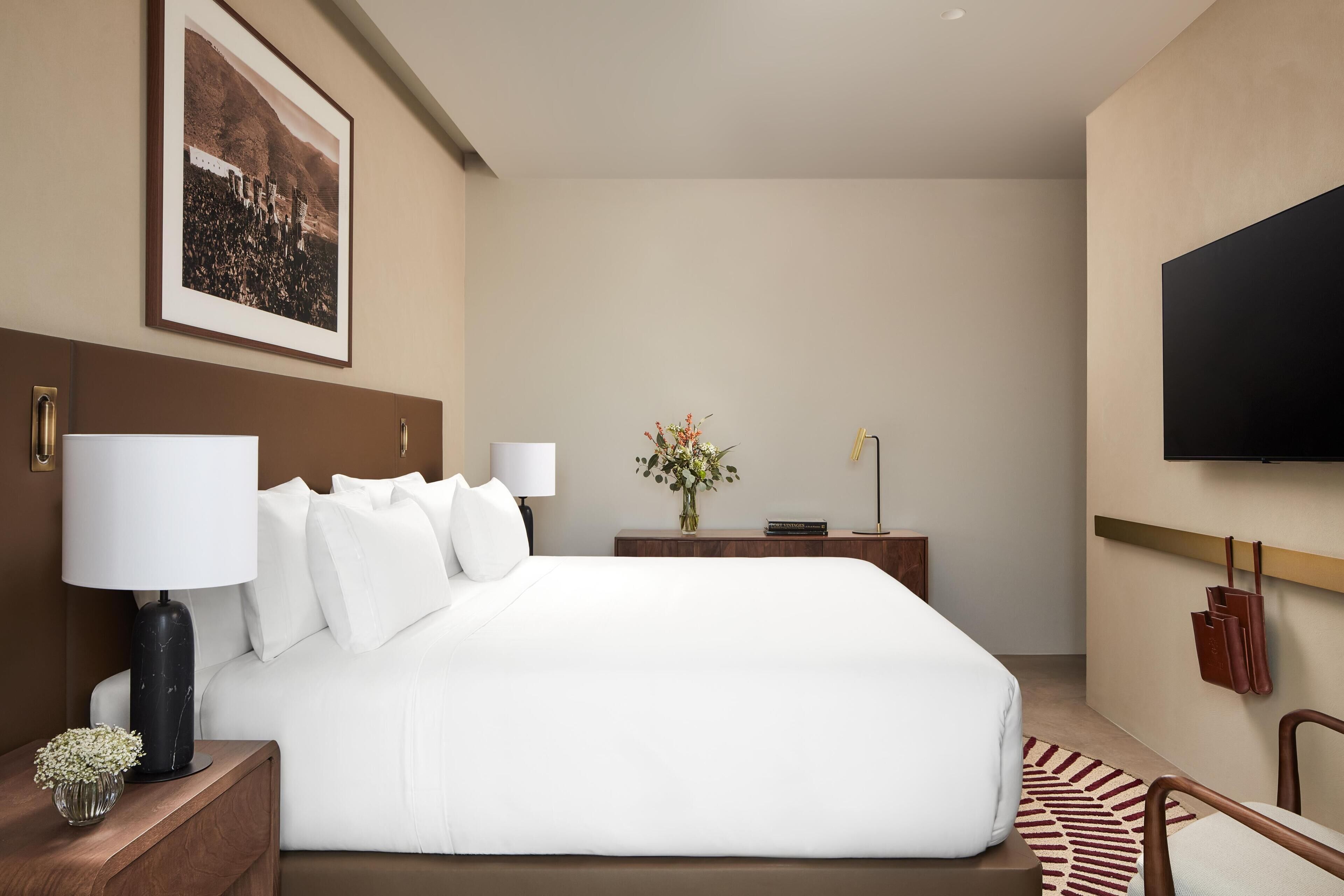A hotel room with a large bed, a flat-screen TV, a nightstand with a lamp, a vase with flowers, and a framed picture on the wall.