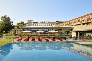Un área de piscina con sillones de descanso y sombrillas, un edificio con un cartel, y un cielo despejado.