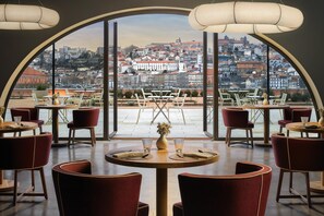 A restaurant with round tables and red chairs, large windows offering a city view.