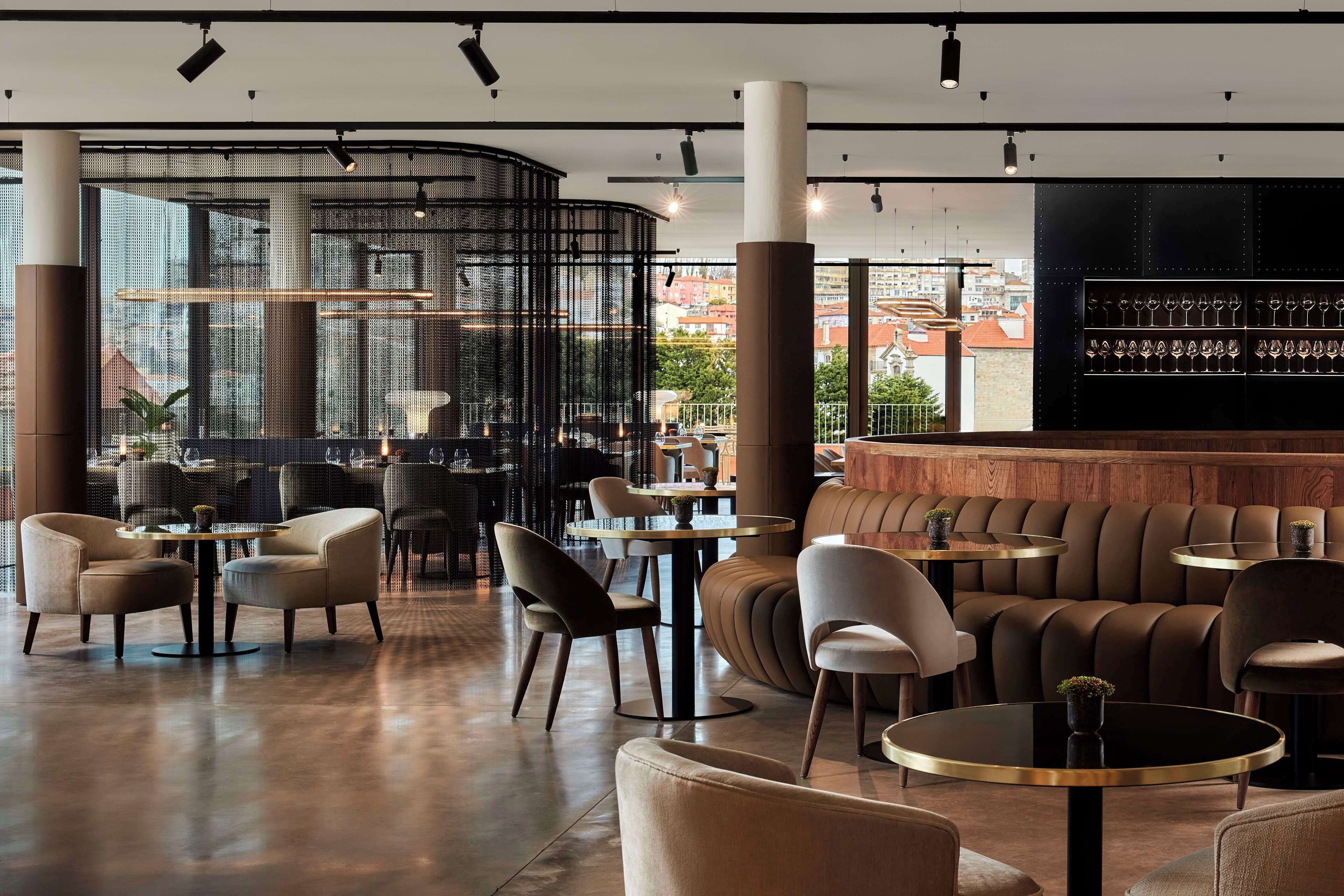 A modern interior with round tables and chairs, a bar area, and large windows offering city views.