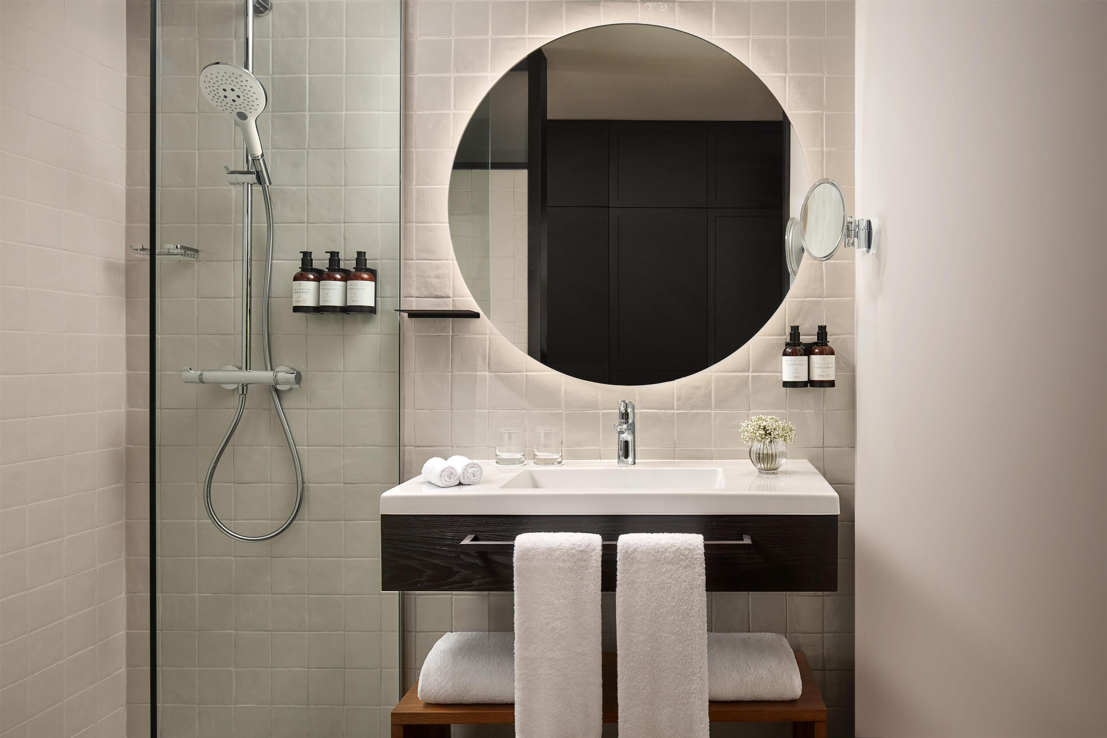 Un baño moderno con ducha envuelta en vidrio, un espejo redondo, un lavabo blanco y un banco de madera.