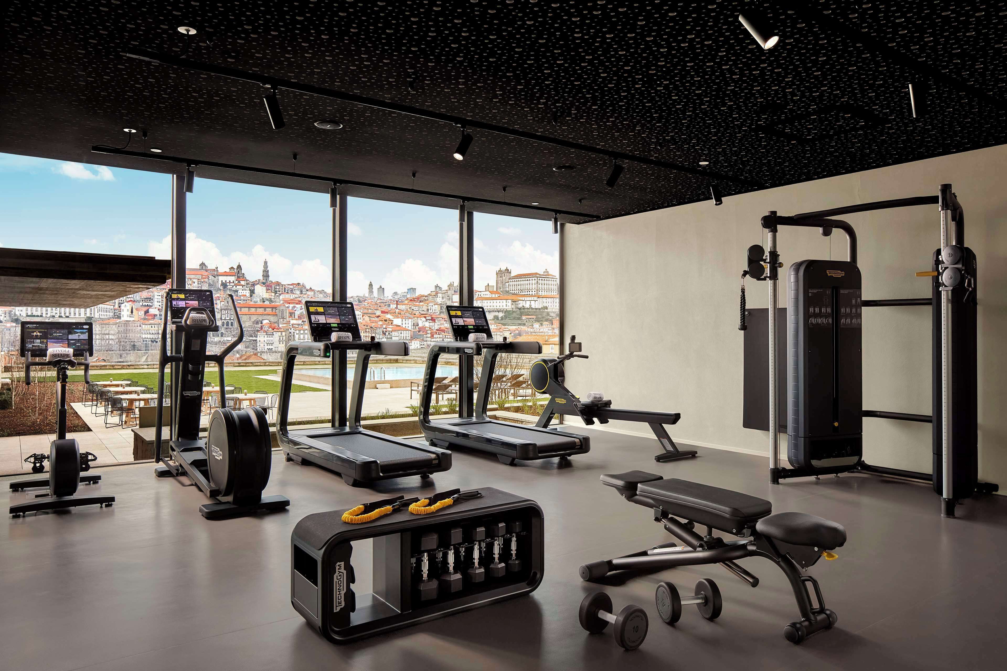 A gym with treadmills, elliptical machines, a bench, dumbbells, and a weight machine, with a city view through large windows.