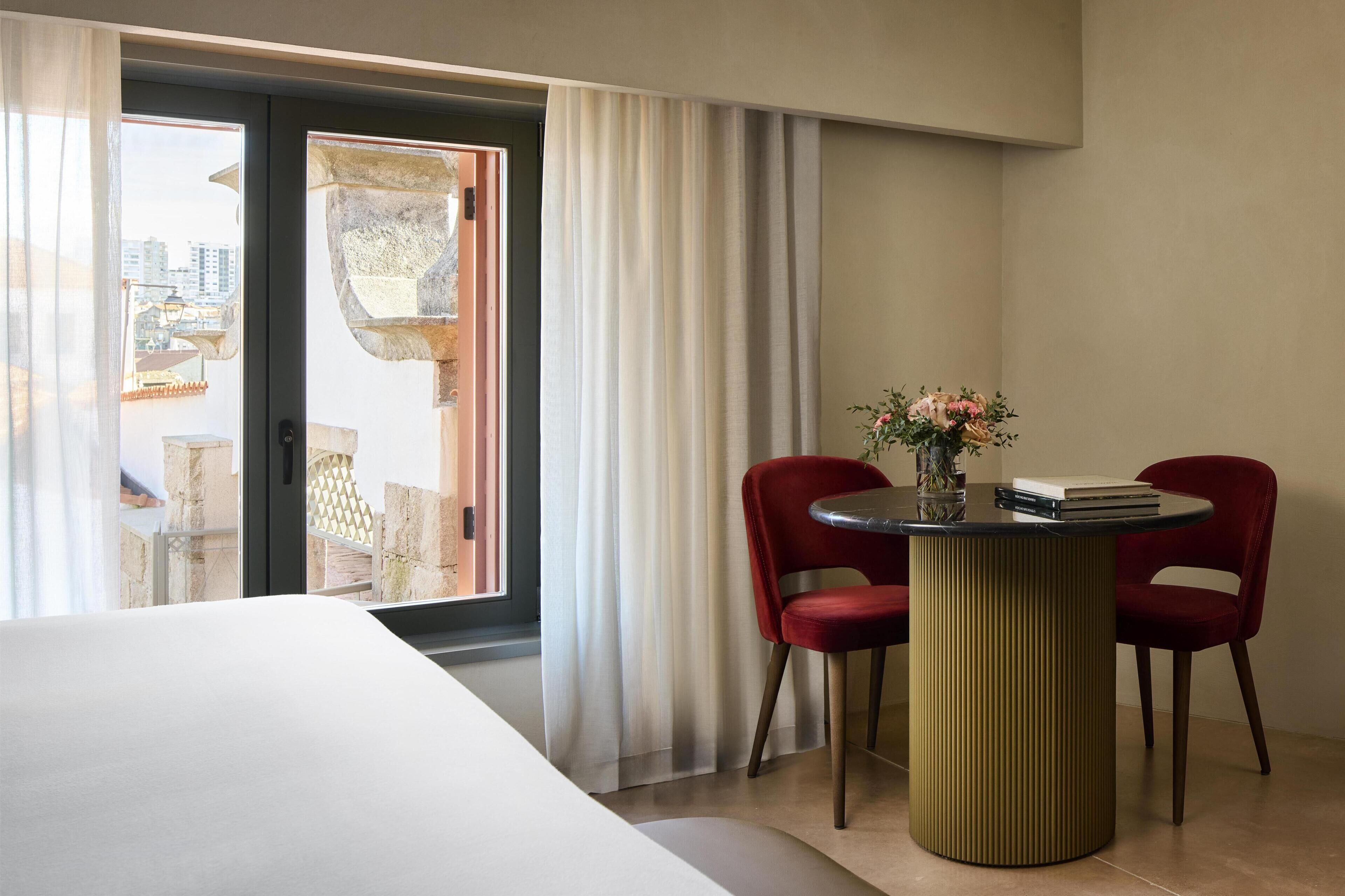 A hotel room with a bed, a table with two chairs, a vase with flowers, and a view through a sliding door.