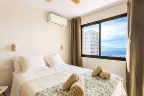 A hotel room with a bed, a window with curtains, an air conditioning unit, and a view of a building and the sea.
