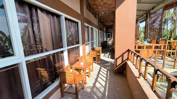 Un espace extérieur abrité, doté de tables et de chaises en bois, de grandes fenêtres et offrant une vue sur la verdure environnante.