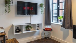 A modern living room with a mounted TV displaying "NETFLIX," a white console table with decorative items, a small side table with a board game, and a window with a view of a brick building.