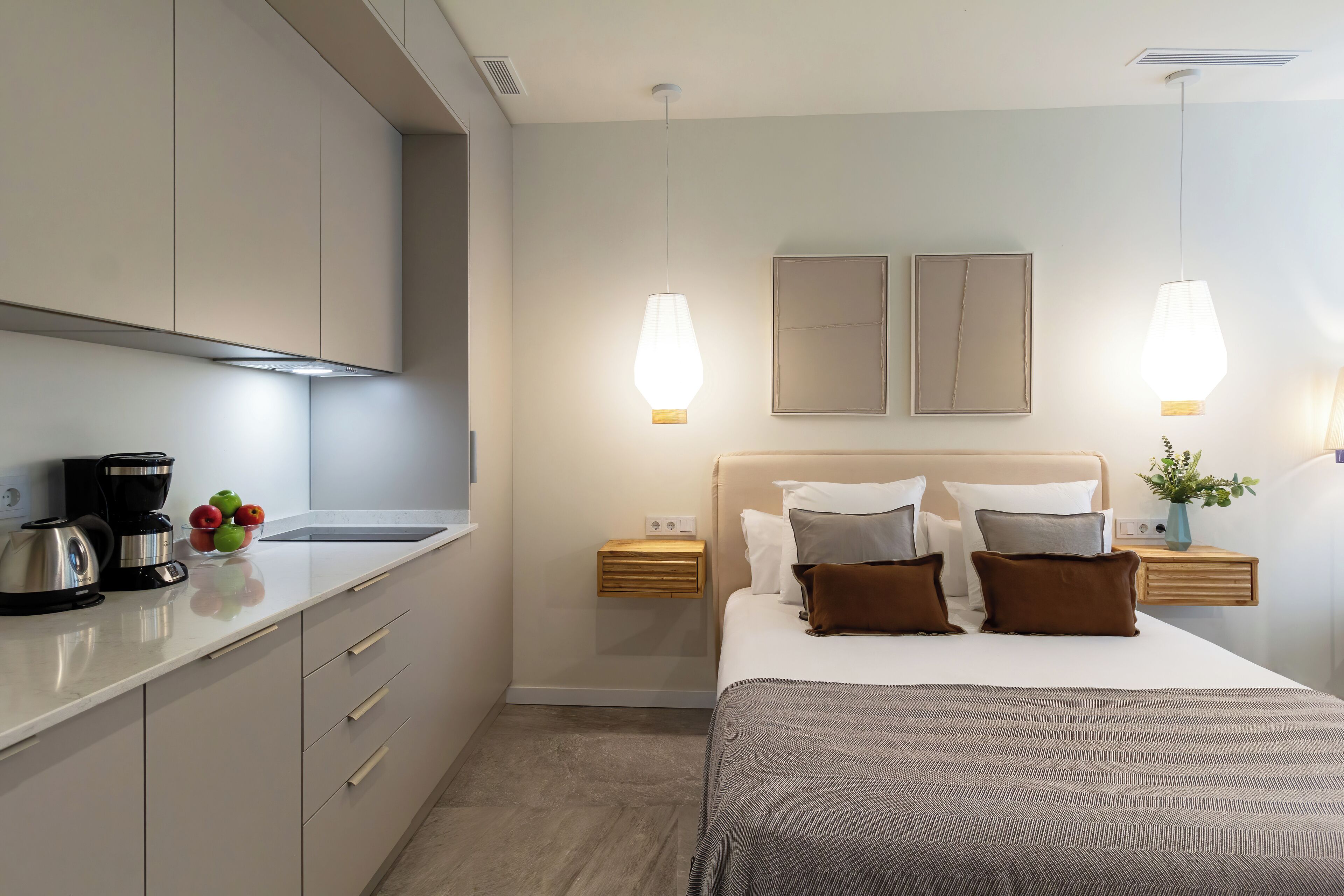A modern hotel room with a kitchenette, a bed with a patterned bedspread, and two bedside tables with lamps.