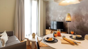 A modern hotel room with a sofa, dining table, breakfast setup, and a television. - Feelathome O'Donnell Apartments (Seville)