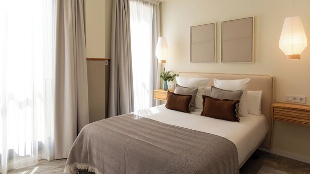 A hotel room with a large bed, two bedside tables, a nightstand, a wall-mounted lamp, and a window with curtains. - Feelathome O'Donnell Apartments (Seville)