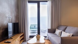 Un salon moderne avec un téléviseur à écran plat posé sur un meuble en bois, une table basse blanche agrémentée d’un vase, un canapé gris et une vue sur l’extérieur grâce à de grandes fenêtres.