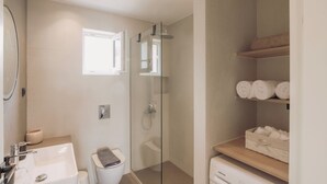 Et moderne badeværelse med en bruseniche med glasvægge, en hvid vask og et toilet. Der er hylder med sammenrullede håndklæder og en lille skraldespand.