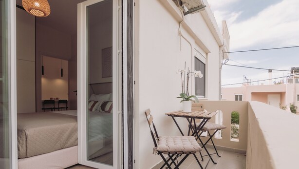 En balkon med et bord og stole, en potteplante og en skydedør, der fører til et soveværelse med en seng og et skrivebord.