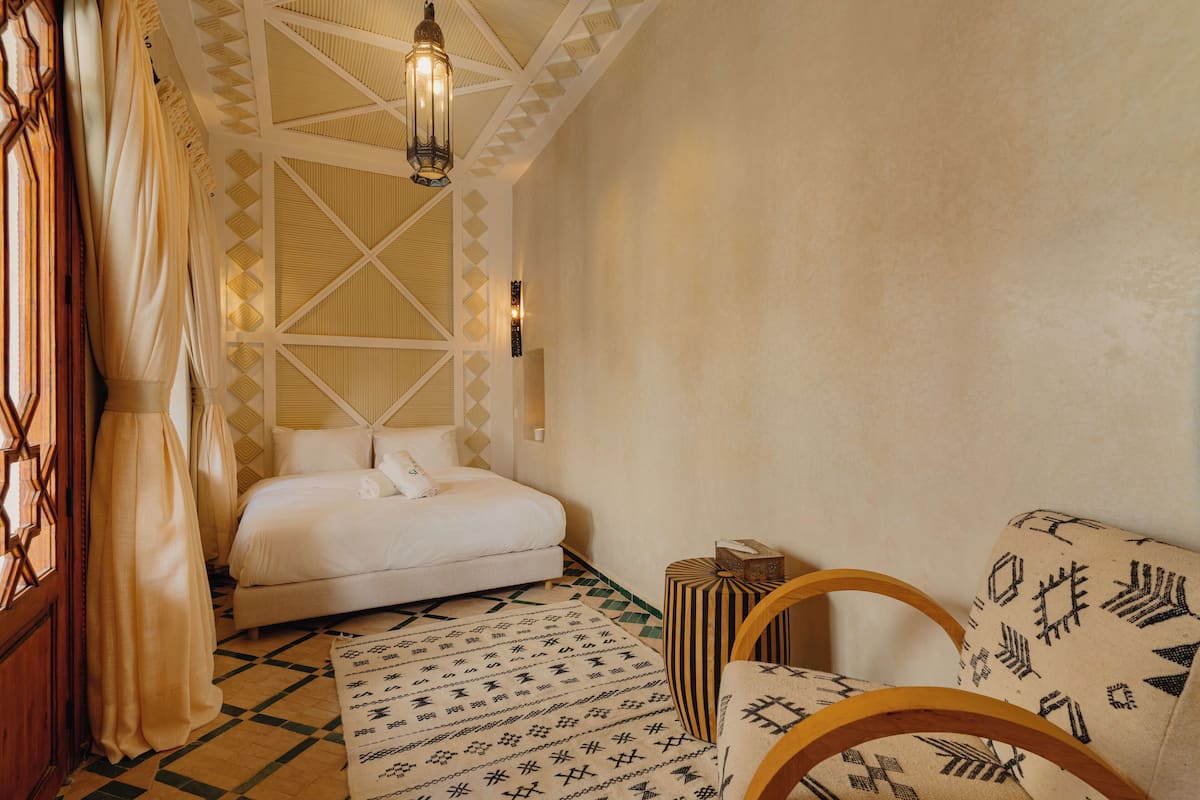 A bedroom with a bed, a patterned rug, a chair, and a hanging light fixture.