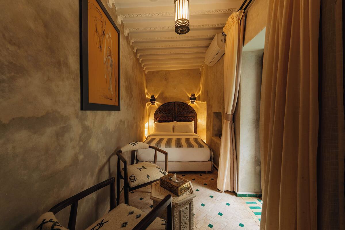 A narrow hallway leading to a bed with a patterned headboard, a chair with a cushion, and a small table with a box.