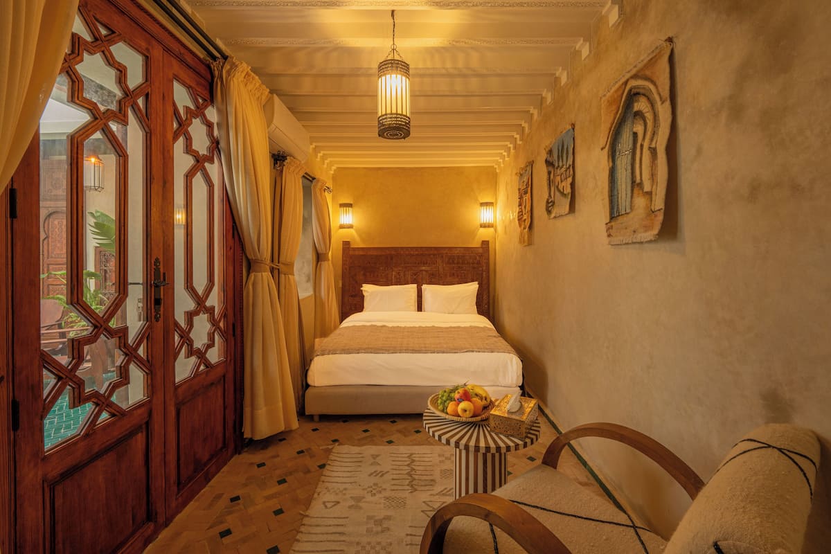 A hotel room with a large bed, a small table with fruit, a chair, and a decorative wall.