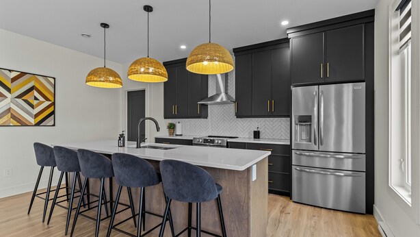 A modern kitchen with dark cabinets, a stainless steel refrigerator, a large island with bar stools, and pendant lighting. - NEW Golfers Dream Modern Patio King Bed (St. John's)