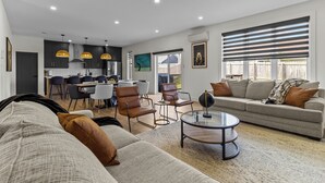 A modern living room with a sofa, armchairs, and a coffee table. There is a dining area with a table and chairs, and a kitchen in the background. - NEW Golfers Dream Modern Patio King Bed (St. John's)
