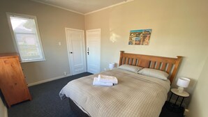 A bedroom with a bed, a wooden headboard, a nightstand with a lamp, a dresser, and a window with blinds. - Close to Trade Central Mall, 3 Bedrooms, Sleeps 6, Wheelchair friendly (Rotorua)