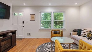 A living room with a sofa, a coffee table, a TV mounted on the wall, and a window with blinds.