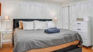 A neatly made bed with a grey and brown bedspread, two bedside lamps, a wooden nightstand, and a white wardrobe with drawers.